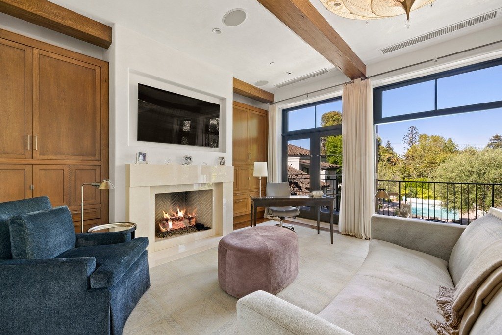 Elegant living area with a fireplace and mounted TV, blue armchair, and desk near large windows overlooking greenery and a pool. Features built-in cabinets, exposed ceiling beams, a light sofa with throw, and a pink ottoman.