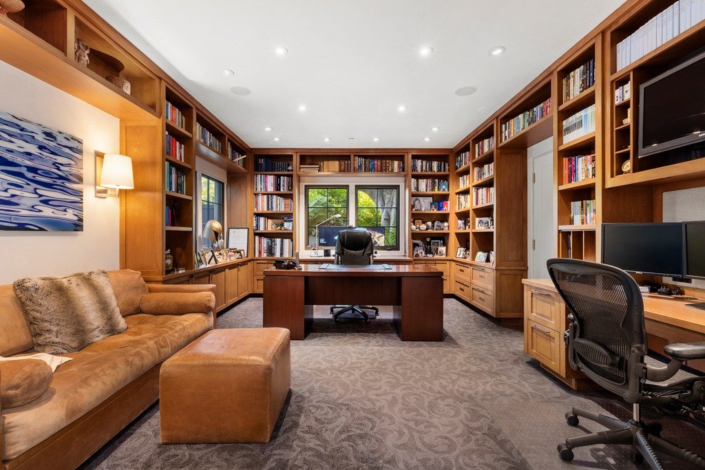 Sophisticated home office with floor-to-ceiling wooden bookshelves, a central dark wood desk facing a window, and a brown leather sofa with ottoman to the side. The room features recessed ceiling lighting, a patterned carpet, and an additional workstation with computer monitors along the right wall.