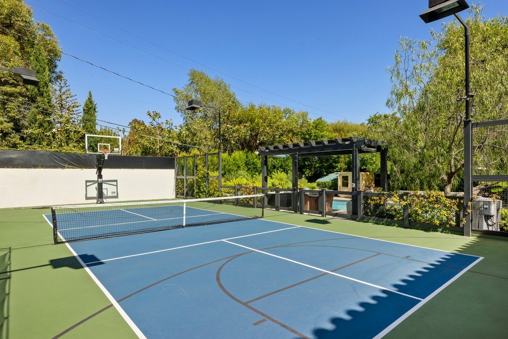 An outdoor blue and green multi-sport court with lines for tennis, pickleball, and basketball, surrounded by trees, with a shaded seating area and a pool visible in the background.