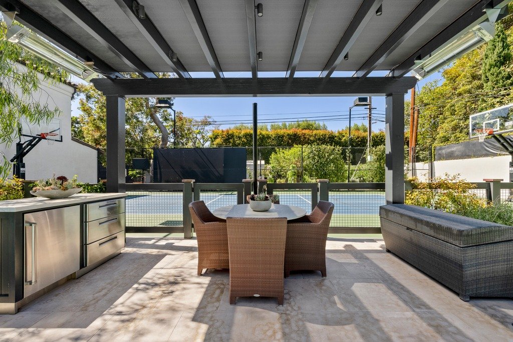 An outdoor patio with a dining table and chairs under a pergola, featuring a kitchen counter with stainless steel cabinets and a wicker storage bench, overlooking a blue tennis court with a basketball hoop.