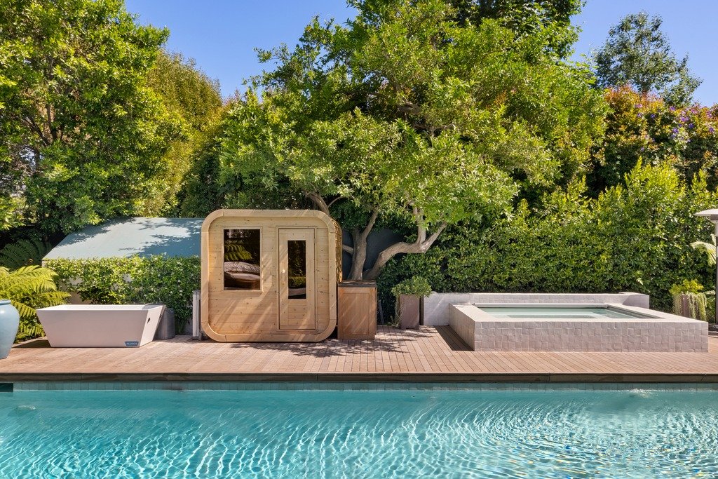 A luxurious outdoor wellness area with a wooden barrel sauna, a modern white cold plunge tub, and a tiled hot tub surrounding a swimming pool on a wooden deck, set against lush green trees.