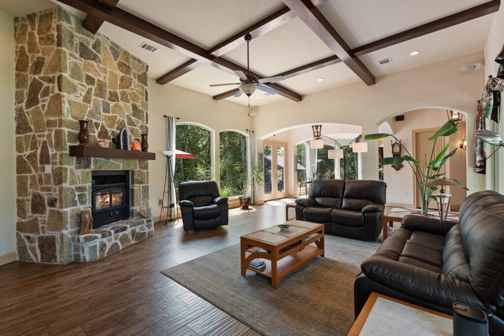 Spacious living room with a stone fireplace and wooden mantle, black leather seating around a wooden coffee table on a light rug, high ceilings with exposed wooden beams, hardwood and tile floors, and large windows