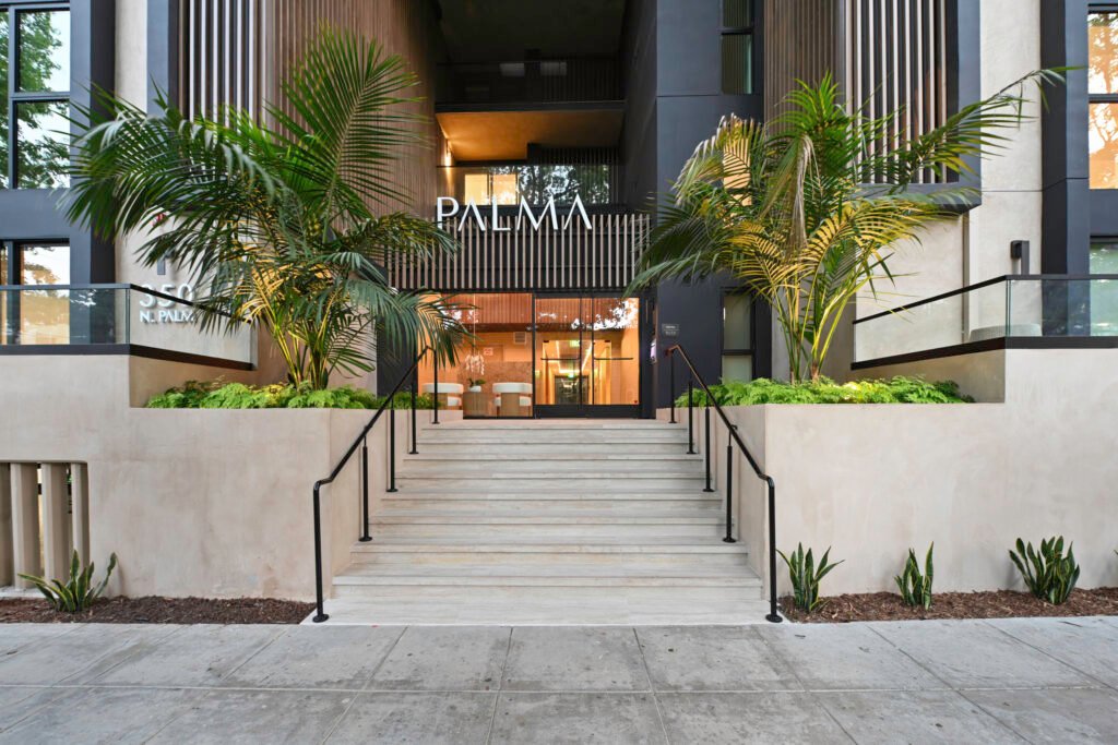 Entrance of Palma Beverly Hills luxury apartments with a wide staircase leading to glass doors, flanked by lush Areca palm trees and manicured planters, and 'PALMA' displayed above the entryway.