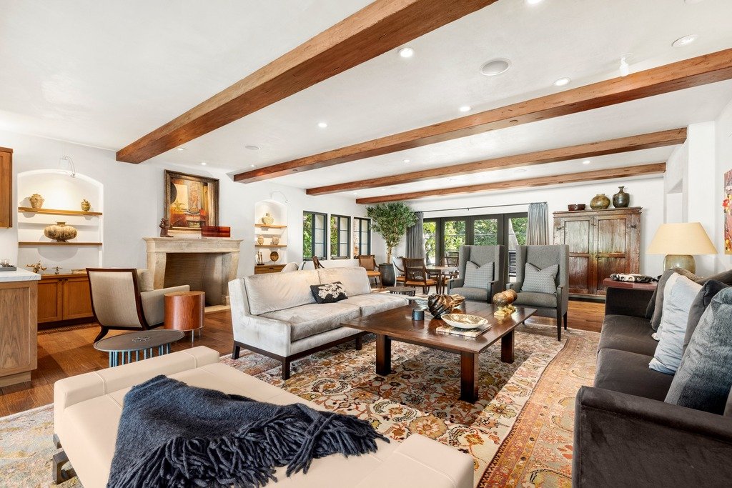 Spacious living room with exposed wooden ceiling beams, patterned area rug, modern and rustic furniture including a long sofa, armchairs, and wooden coffee table, with large windows framing views of a dining area and outdoor space.