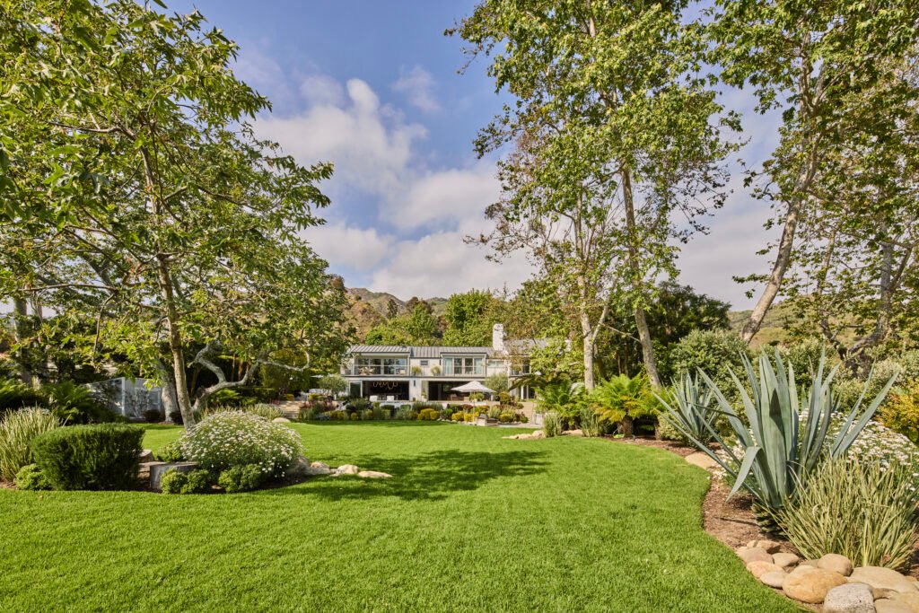 A large, well-kept lawn leading to a modern house with multiple windows and balconies, surrounded by lush greenery and landscaping under a partly cloudy Malibu sky.