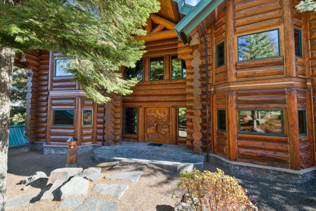 A large multi-level log cabin with a green roof and many windows, set on a hillside with a stone retaining wall, surrounded by tall pine trees, featuring a deck with outdoor furniture overlooking the forest and distant mountains.