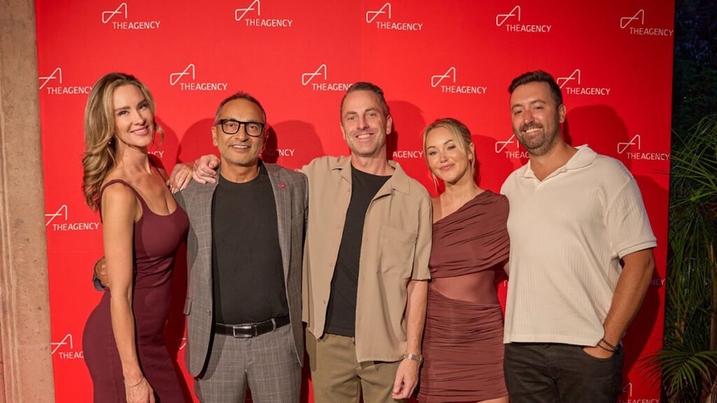 A group of five people, three men and two women, stand together smiling in front of a red wall with "The Agency" logo repeated on it. From left to right: a woman with long, blonde hair wearing a form-fitting burgundy dress; a man with graying hair, a beard, and glasses wearing a checkered suit jacket over a black shirt; a man with a shaved head and a beard wearing a beige button-down shirt; a woman with long, blonde hair wearing a one-shoulder brown dress; and a man with a beard and short, dark hair wearing a white polo shirt.