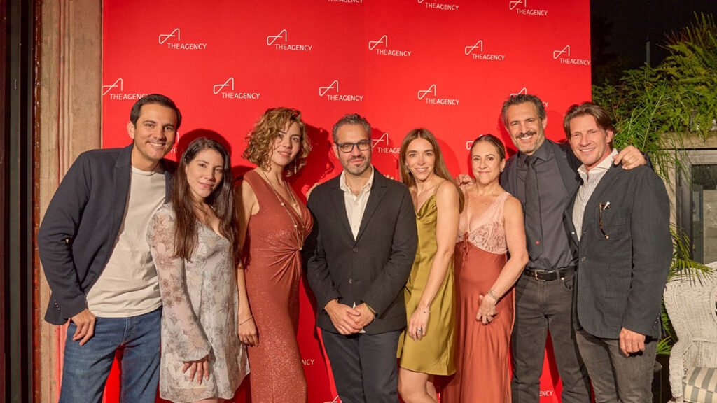 A group of seven people—four men and three women—stands together, smiling, in front of a red wall patterned with The Agency logo. The people are dressed in a mix of casual and formal attire
