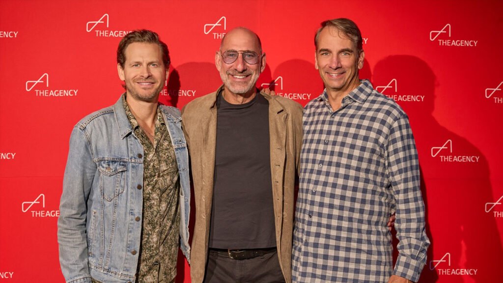 A medium shot of three men standing side-by-side in front of a red wall with The Agency logo. From left to right: a man with a beard and light brown hair wearing a denim jacket over a patterned shirt; a bald man with a beard and sunglasses wearing a beige jacket over a dark t-shirt; and a man with graying hair and a beard wearing a blue and white plaid shirt.