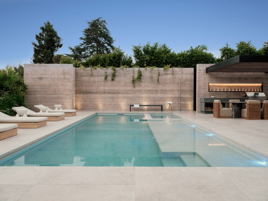 Modern architectural estate featuring a sparkling swimming pool, spacious paved patio with multiple lounge chairs, and an outdoor kitchen/bar area, all enclosed by a minimalist concrete wall and lush landscaping.