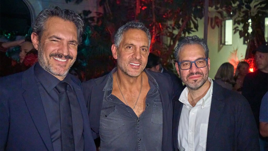 A medium shot of three men standing side-by-side outdoors at night, smiling at the camera.