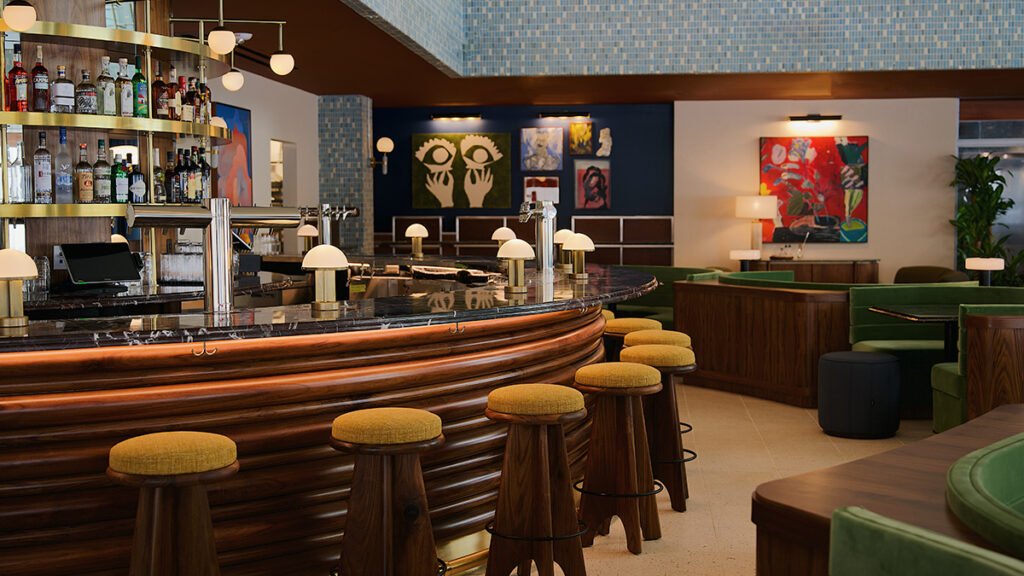 Mid-century modern hotel lobby bar with a curved wooden counter, and green velvet banquettes.