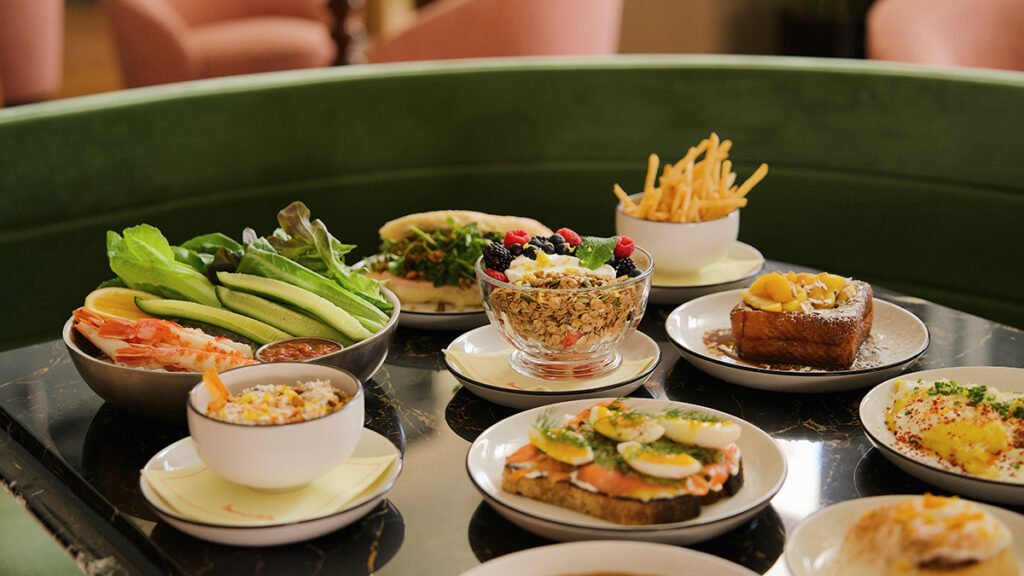 A table filled with a gourmet brunch spread, including a yogurt parfait, smoked salmon toast, french toast, shrimp salad, and fries