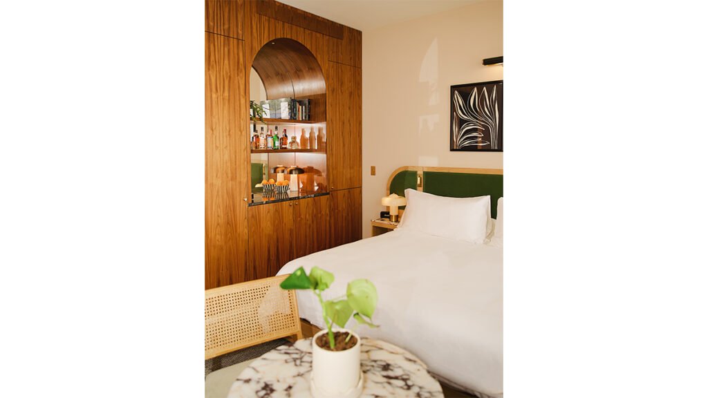 A mid-century modern hotel room with a green velvet headboard, a large wood-paneled minibar, and a white bed.