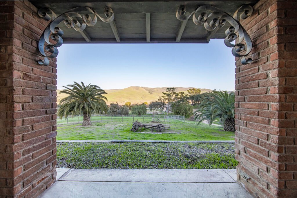A green field with trees and hills seen through a red brick archway with decorative metalwork; palm trees and old farm equipment are visible.