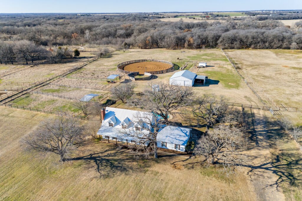 Aerial view of a rural property with a house, barn, horse training ring, and open fields surrounded by bare trees.