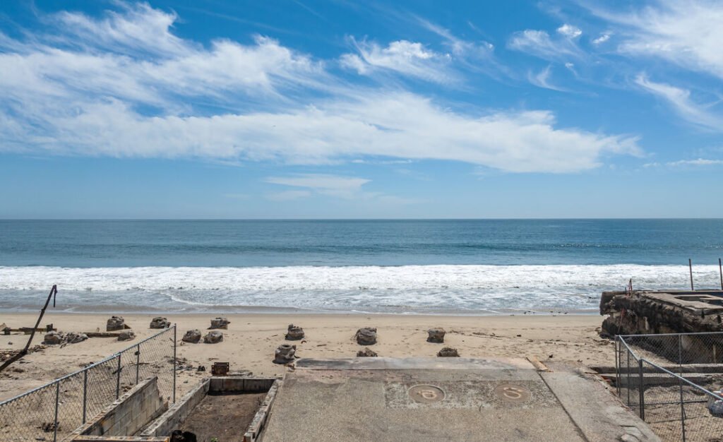 Remains of a structure on La Costa Beach in Malibu, CA, with concrete foundations, debris, and fencing on a sandy beach, the ocean in the background, and a clear blue sky.