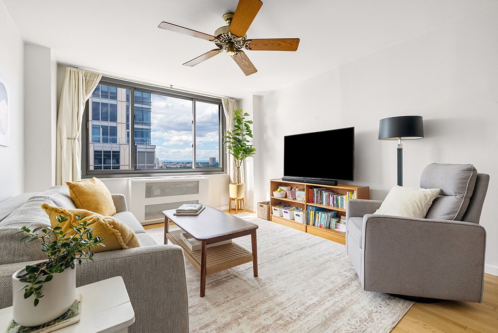 Modern living room with grey sofa and armchair, wooden coffee table, and a large window overlooking the city.