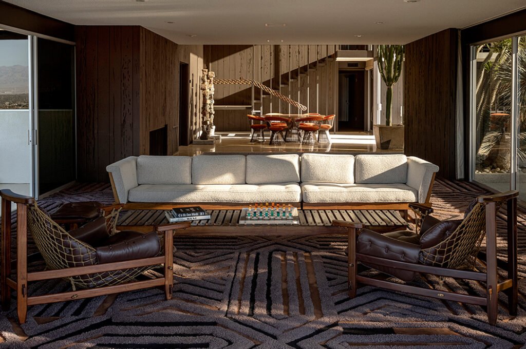 A mid-century modern living room with wood paneling, a large light gray sofa, a wood slatted coffee table, and two brown leather and rope lounge chairs, with a dining area and staircase in the background.