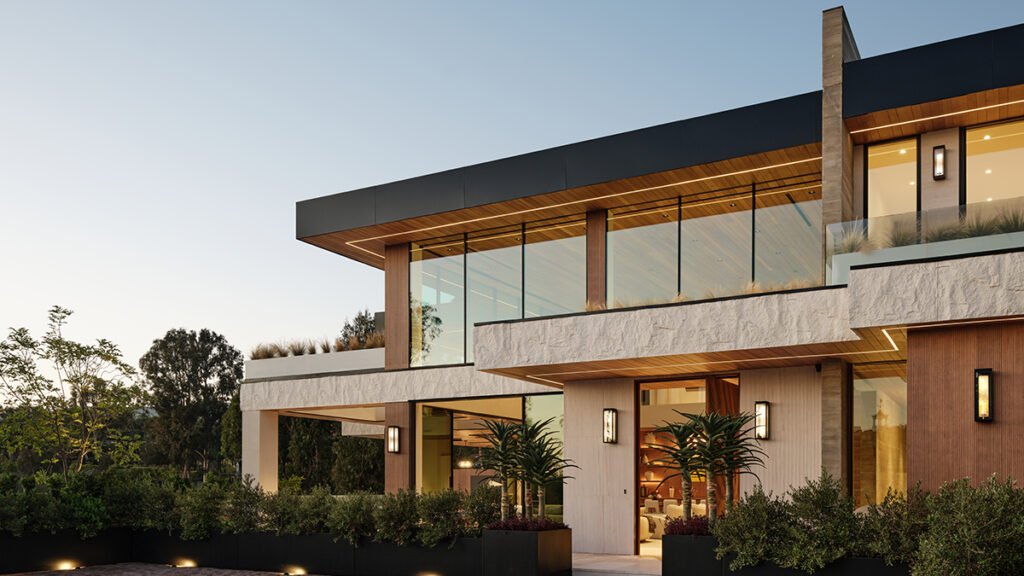 Facade of a modern luxury estate at dusk, featuring contemporary architecture with extensive floor-to-ceiling glass walls, integrated lighting, wood accents, hand-chiseled Portuguese limestone, and lush landscaping.