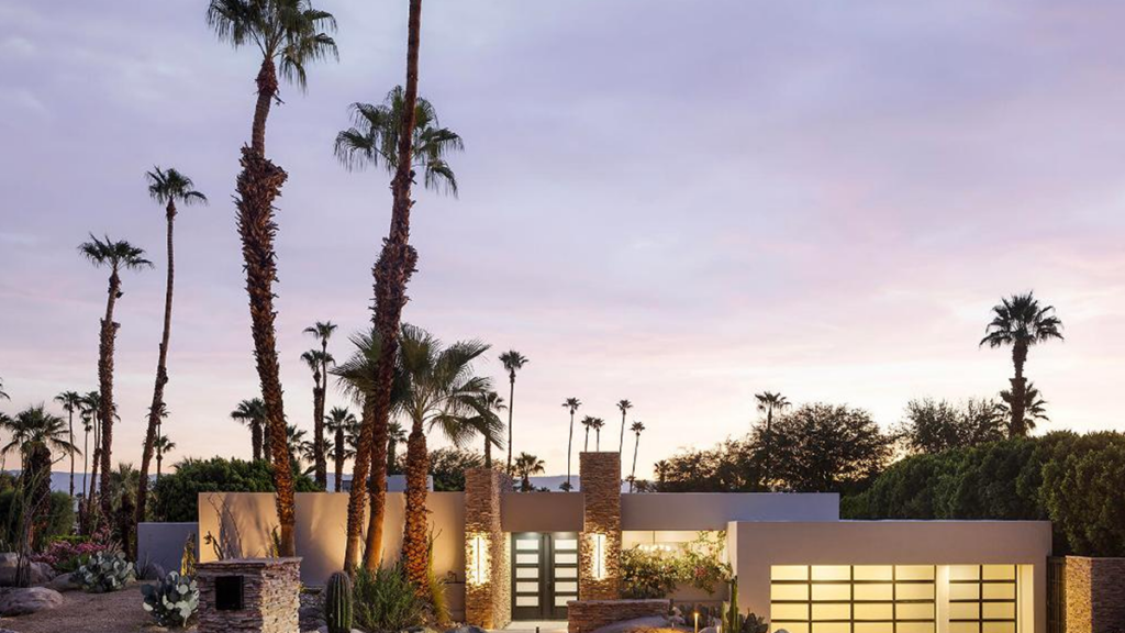 A striking single-story, beige modern desert home illuminated at dusk, featuring a stone entryway and two glass garage doors, surrounded by tall palm trees and desert landscaping.