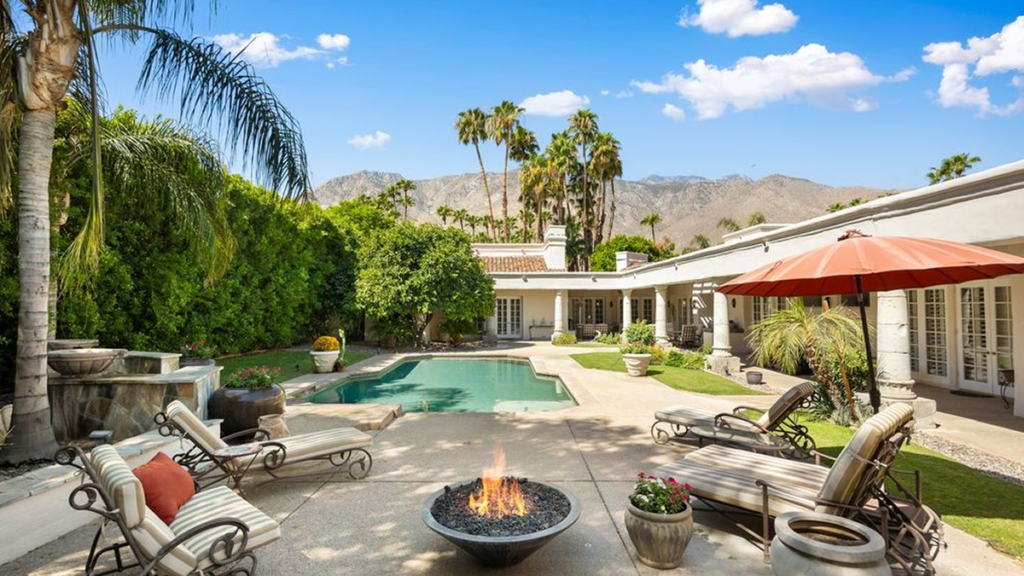 A inviting back patio of a luxury desert home in Palm Springs, featuring a swimming pool, several lounge chairs, a lit fire pit, tall palm trees, and stunning mountain views.