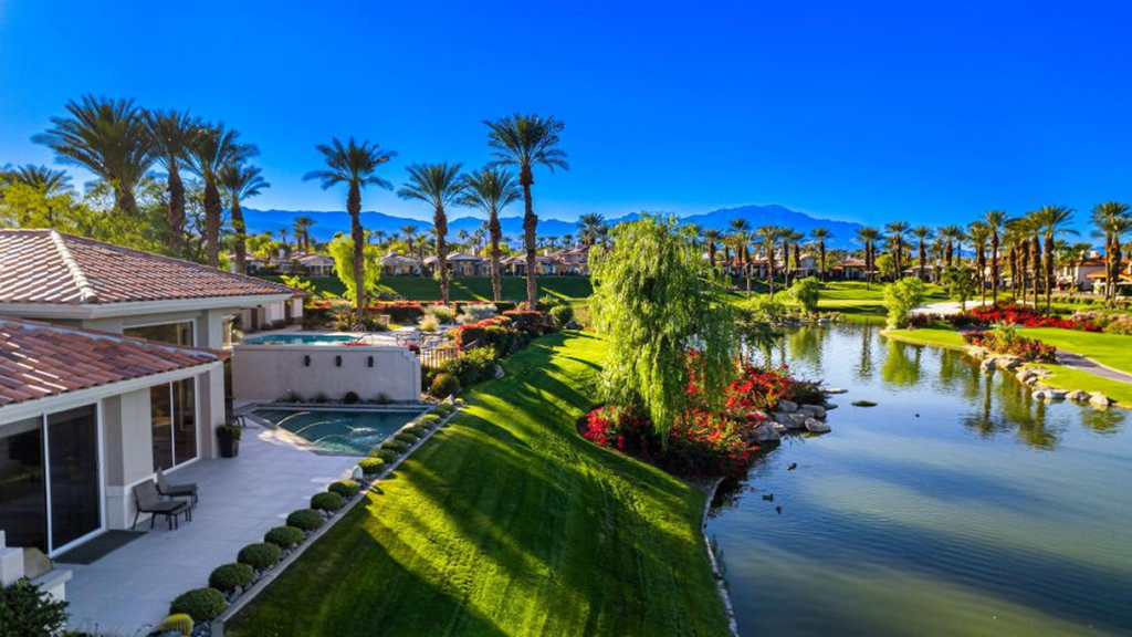 An aerial view of a luxury house with a pool overlooking a lush green golf course, a tranquil lake, tall palm trees, and mountains in the background, likely in Palm Desert, California. 