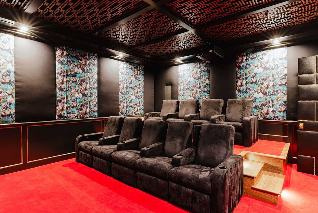 Luxury home cinema at The Eden in Encino featuring velvet reclining theater seats, vibrant red carpeting, and an intricate fretwork ceiling with colorful abstract wall panels.