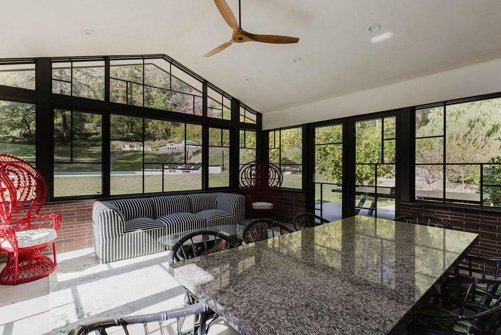 Sunroom at The Eden in Encino featuring Mondrian-inspired window walls, a striped sofa, red peacock chairs, and views of the 2.6-acre botanical grounds.