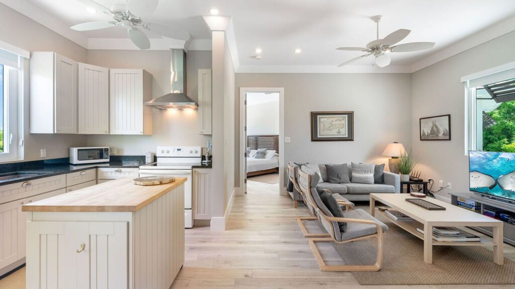 An open-plan coastal living room and kitchen area with light wood floors and white cabinetry.