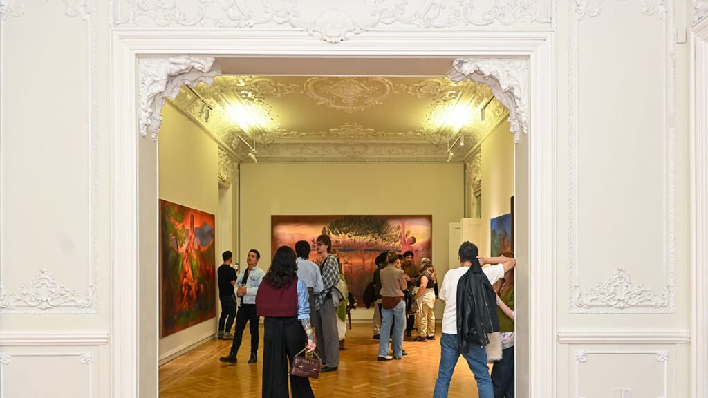 An interior view of a crowded art gallery opening featuring people socializing and viewing contemporary paintings on a brightly lit wall.