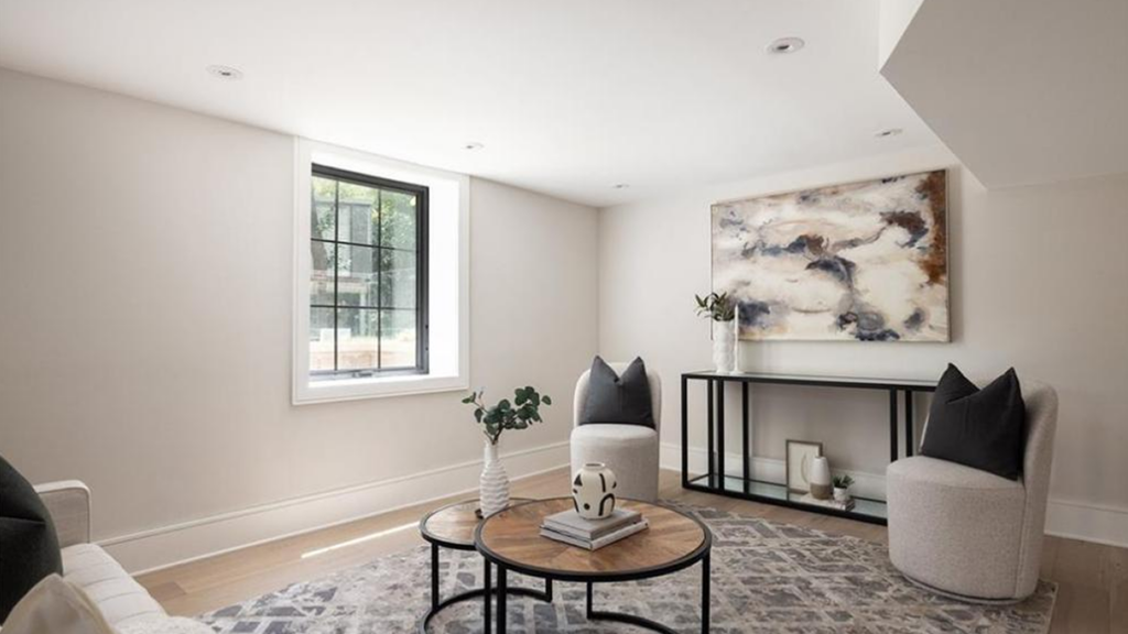 Modern basement sitting area with black-framed grid window, abstract art, grey swivel chairs, and round wood coffee tables.