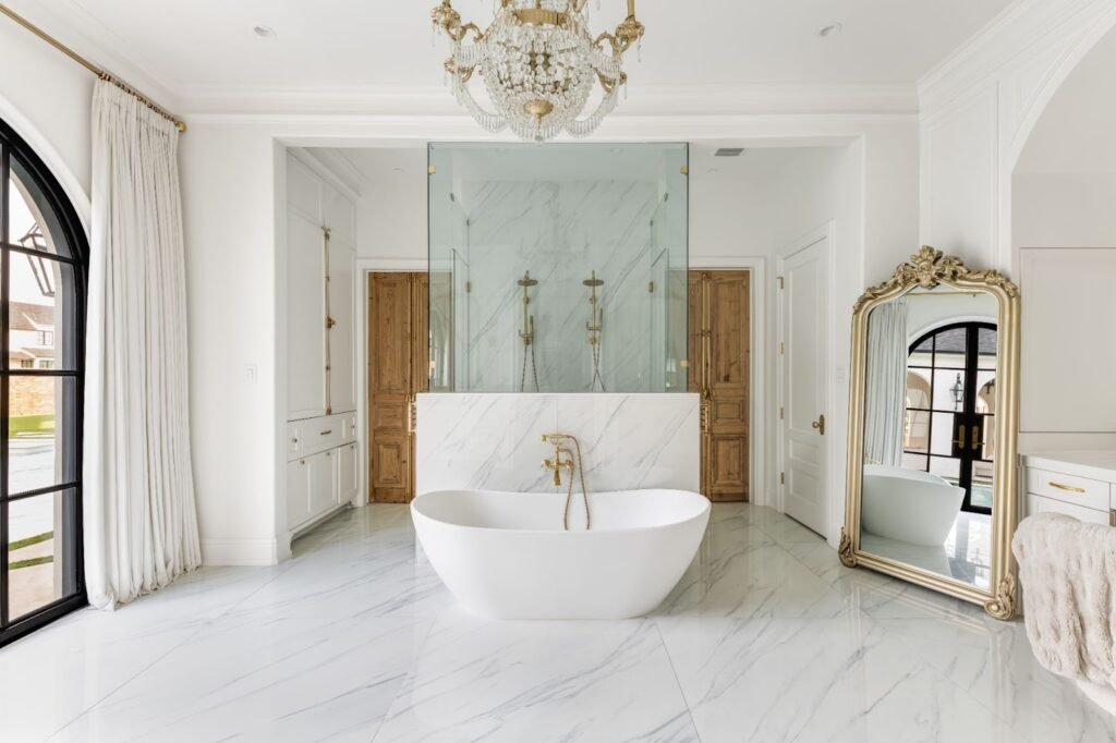 Luxury primary bathroom in Frisco, TX with a freestanding tub, marble floors, a crystal chandelier, and a gold floor mirror.