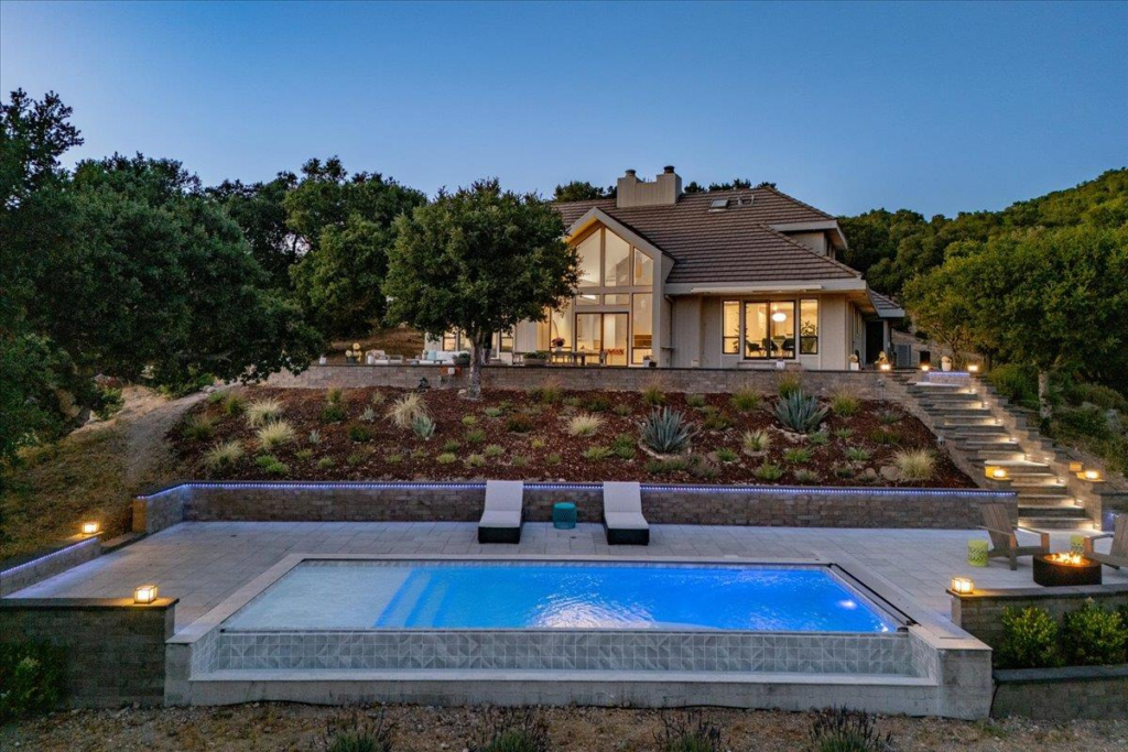 Dusk view of the Via Las Rosas retreat in Carmel Valley featuring a tiered patio, illuminated pool, and modern architecture.