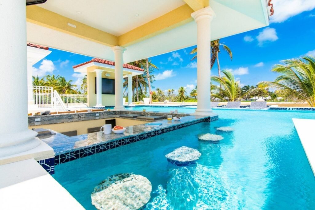 Luxury swim-up pool bar at Villa Del Mare, Grand Cayman, featuring granite countertops, submerged bar stools, and a shaded poolside pavilion with classical white columns.