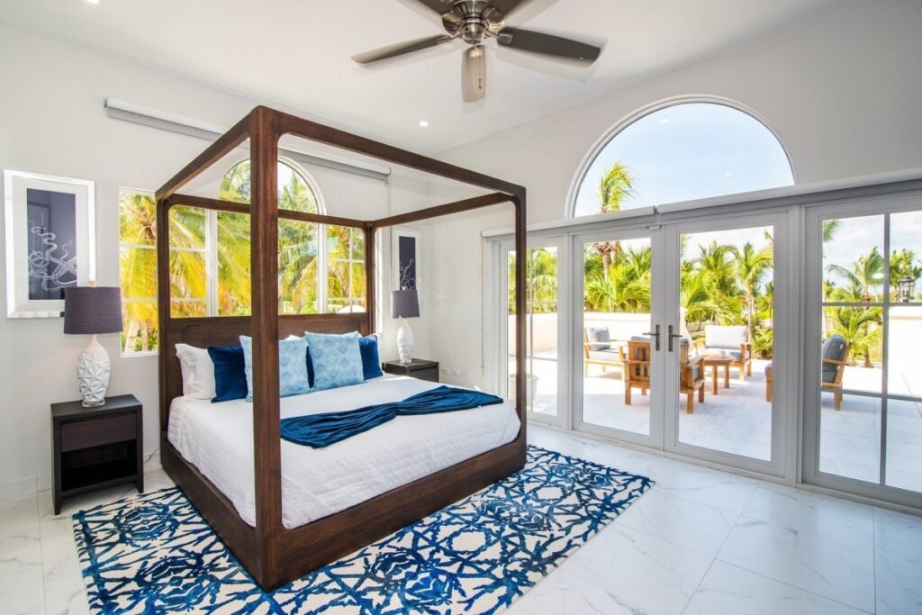 Luxury primary bedroom at Villa Del Mare in Grand Cayman, featuring a solid wood four-poster canopy bed, marble floors, and arched French doors opening to a private beachfront terrace with tropical garden views.