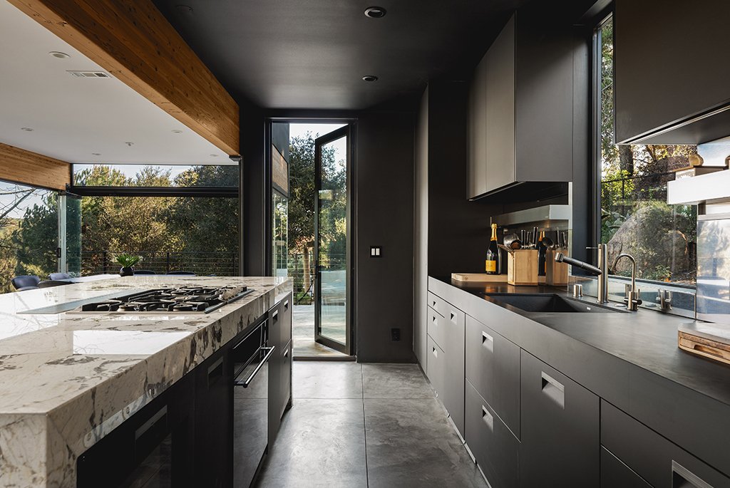 Modern Arclinea kitchen in a Malibu home by Finn Kappe, featuring a sculptural marble island with a gas range, dark minimalist cabinetry, and concrete floors.