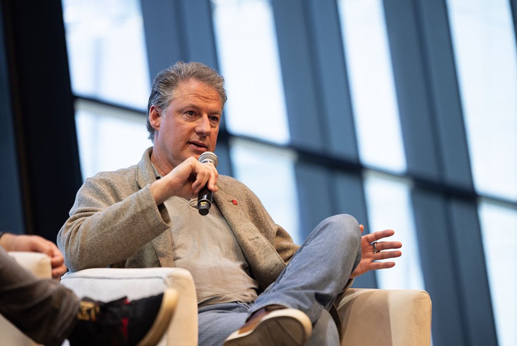 Architect Finn Kappe speaking at a professional panel discussion, wearing a grey blazer and red lapel pin against a modern city skyline backdrop.