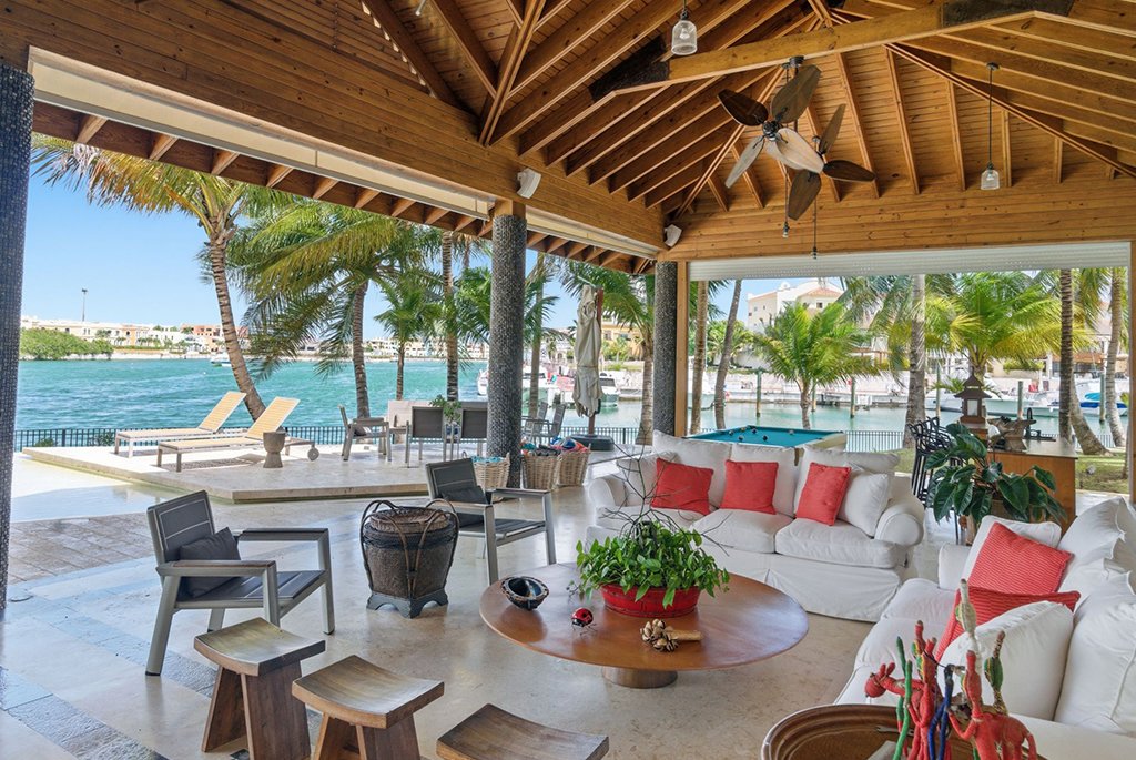 Open-air luxury pavilion lounge with high wooden ceilings, plush white sofas with red accent pillows, and views of a waterfront patio.