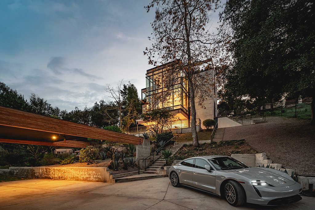 Modern architectural home by Finn Kappe in Malibu, featuring a glass-wrapped exterior, landscaped grounds, and a silver Porsche Taycan parked in the gated driveway at dusk.