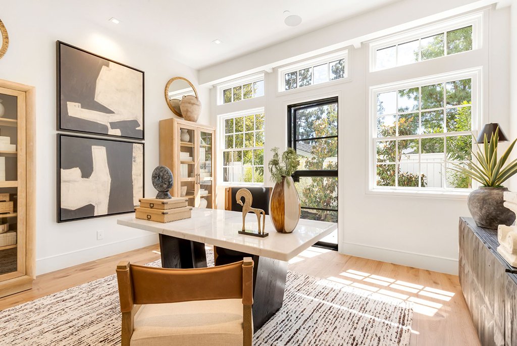 Luxury modern home office featuring a marble executive desk, leather accent chair, large black-frame windows, natural wood display cabinets, abstract wall art, and an external glass door.