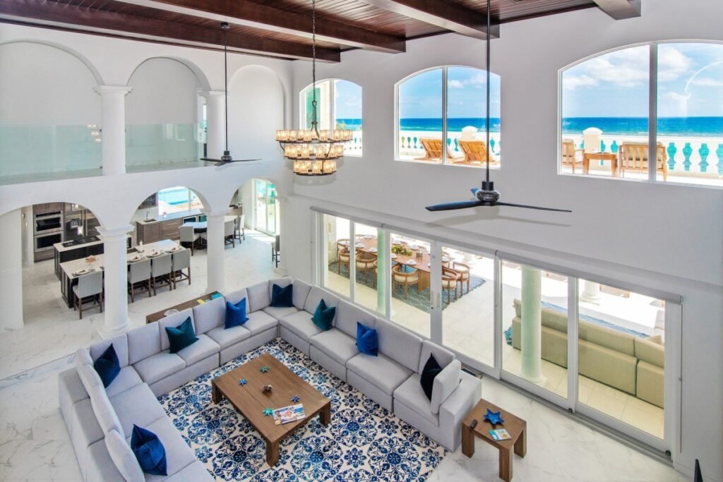 Interior living room of Villa Del Mare in Grand Cayman, featuring a double-height ceiling with exposed dark wood beams, arched windows overlooking the Caribbean Sea, and a large sectional sofa on a blue and white patterned area rug.