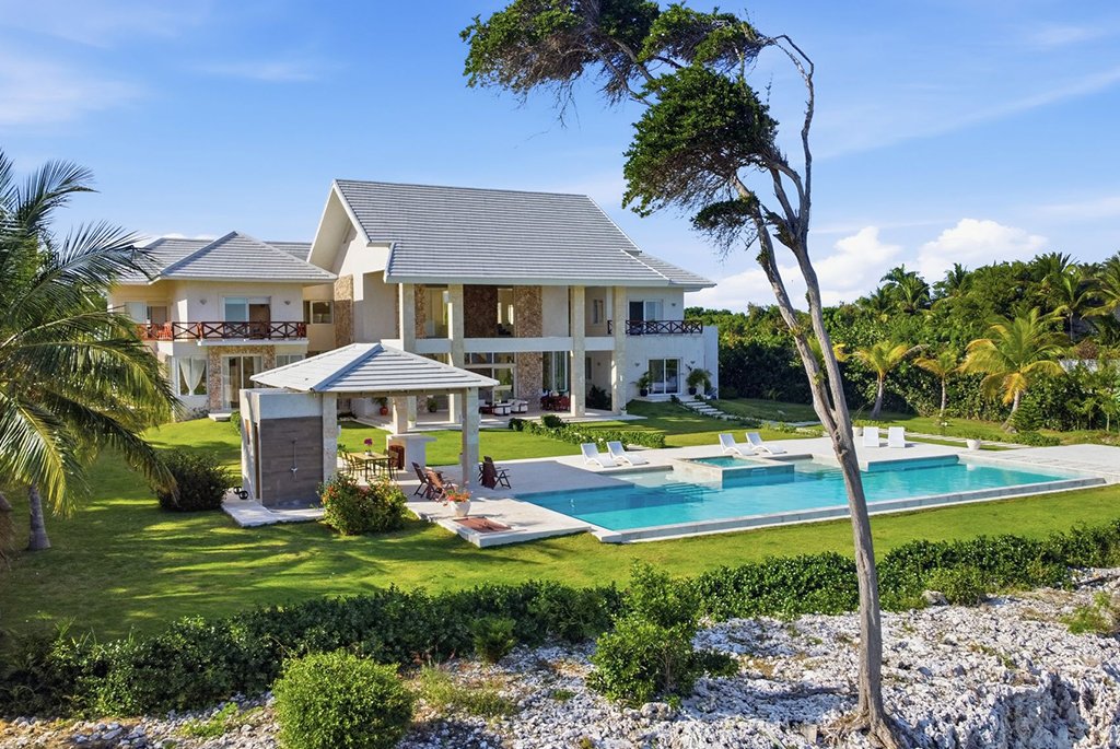 Aerial view of a luxury multi-story oceanfront estate featuring a large swimming pool, open-air gazebo, and manicured green lawn.