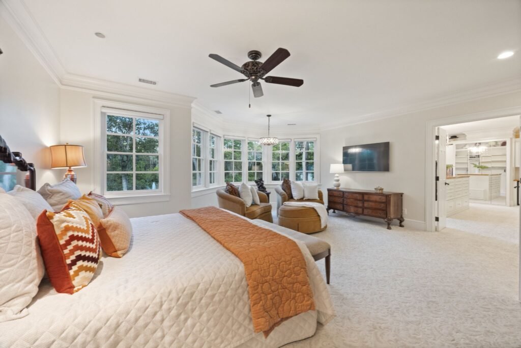 A luxury master bedroom featuring a large bed with orange accents, a sitting area with armchairs, a ceiling fan, and an open doorway leading to a walk-in closet.