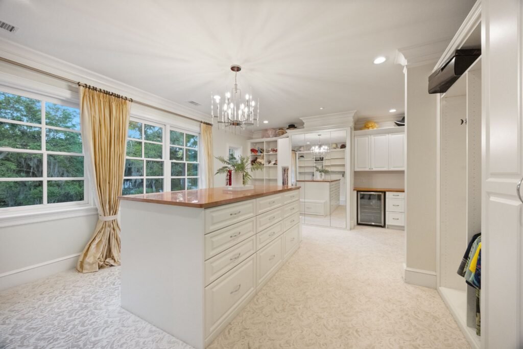 A luxury walk-in closet featuring a large center island with drawers, white custom shelving, a crystal chandelier, and a built-in beverage cooler.