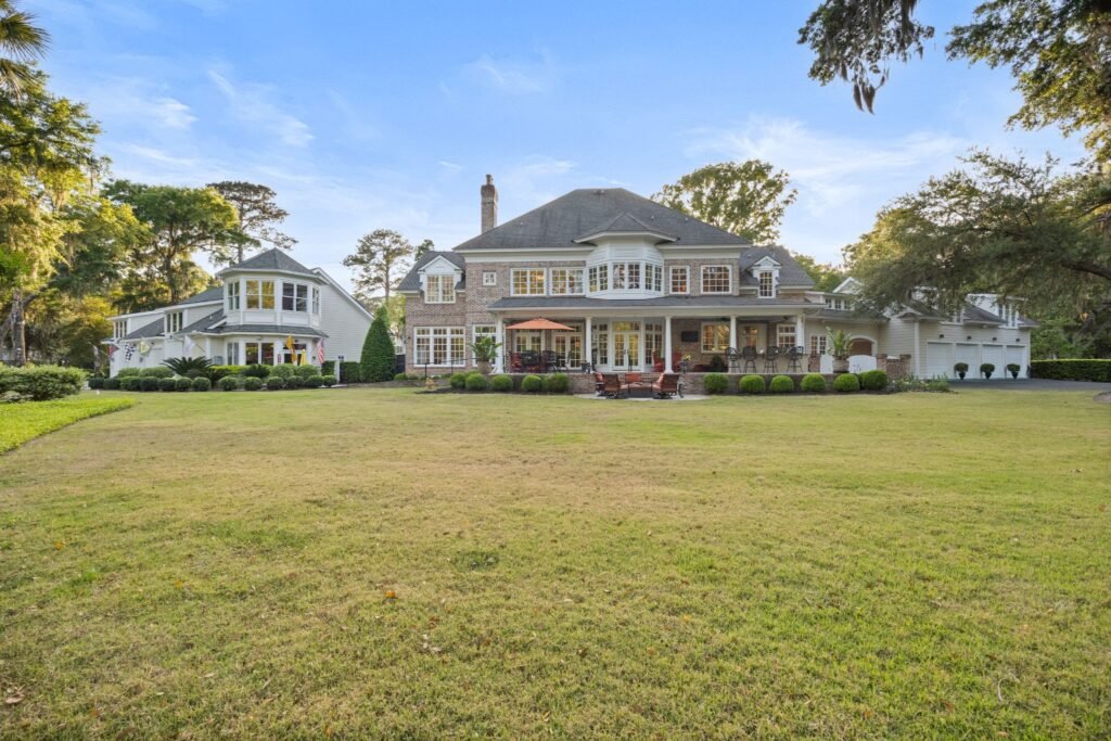 Rear exterior view of a massive luxury brick estate featuring a sprawling green lawn, a covered patio, a separate guest house, and a multi-car garage.