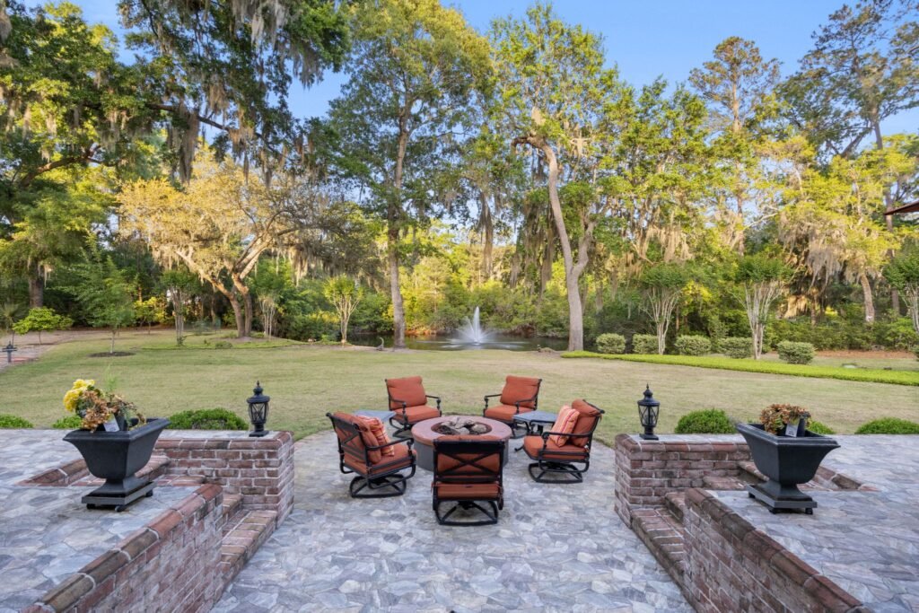 A luxury stone patio featuring an outdoor fire pit with patio chairs overlooking a spacious lawn, mature trees, and a pond with a fountain.