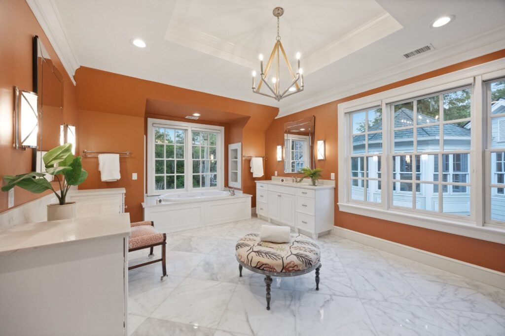 A luxury primary bathroom featuring terracotta orange walls, white double vanities, marble tile flooring, and a central upholstered ottoman under a geometric chandelier.