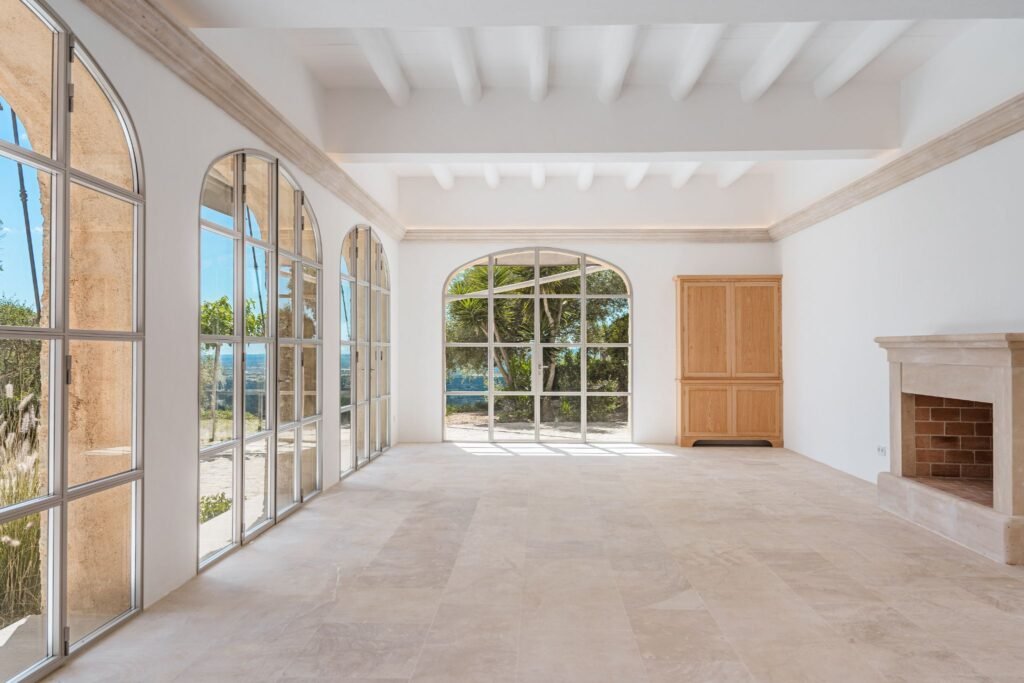 Luxury Mediterranean home interior with large arched floor-to-ceiling windows, white exposed beams, and a stone fireplace.