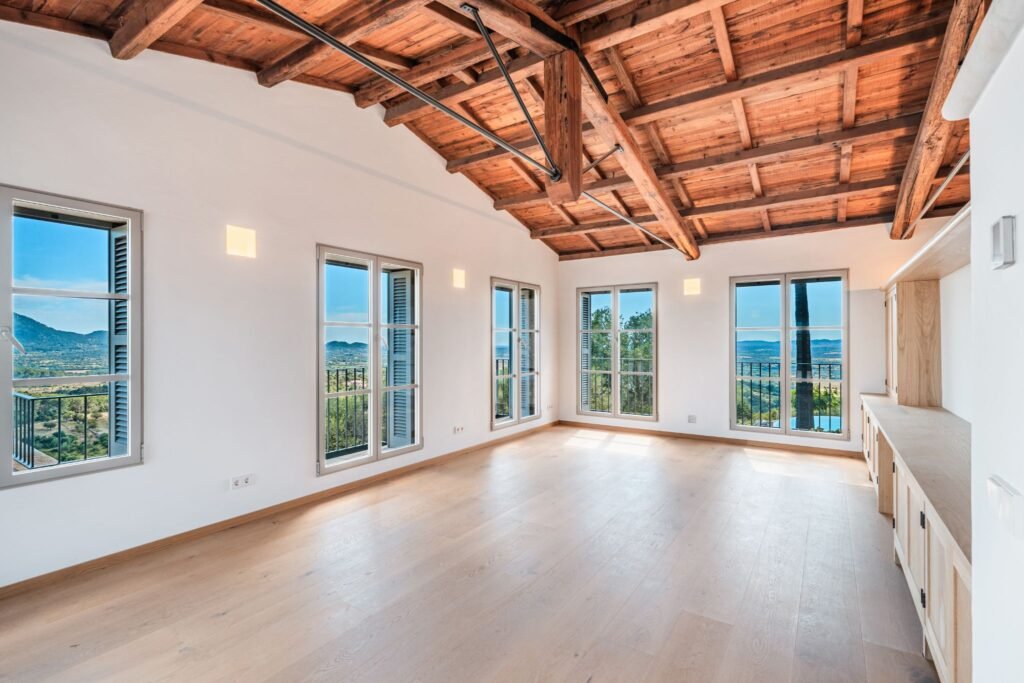 Luxury home interior featuring a vaulted exposed wood beam ceiling, light oak hardwood floors, and multiple windows with panoramic mountain views.